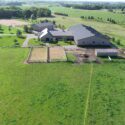 Drone view of full property with large fenced pastures in front, building behind, riding area further back