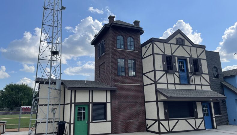Left to right: Short tan building with green door, tall brown brick building, tan building with blue doors